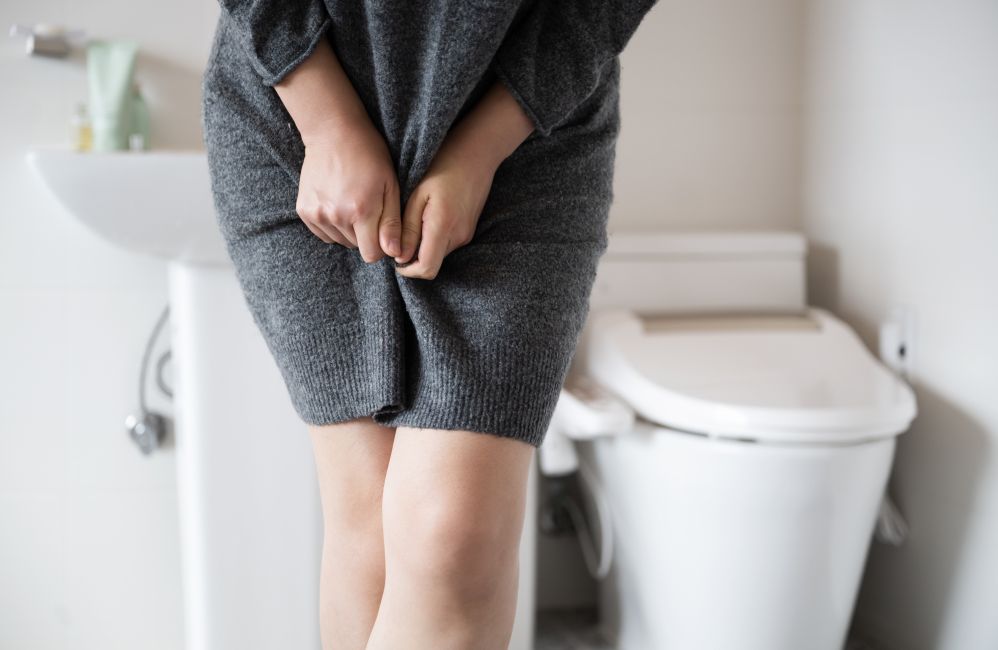 Das Bild zeigt einen Ausschnitt eines Badezimmers mit einer weißen Toilette im Hintergrund und eine Person in einem grauen Strickkleid