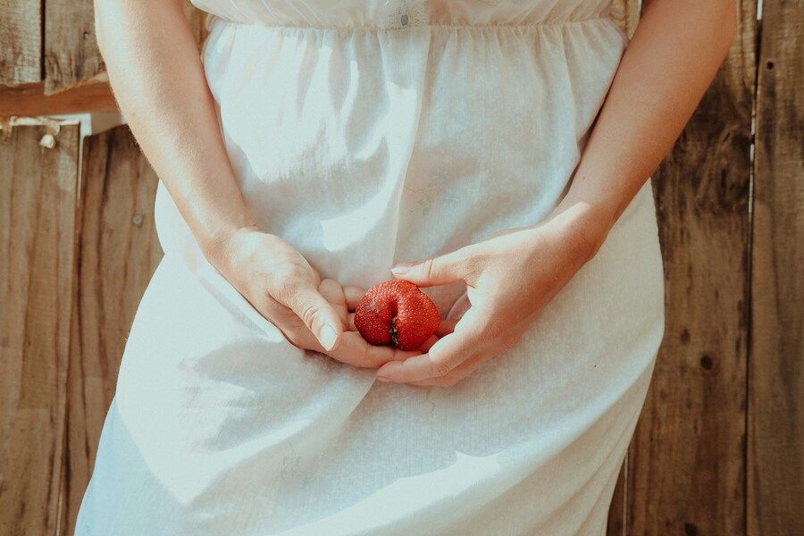 Ein Frau im weißen Kleid hält sich eine große Erdbeere vor den Genitalbereich