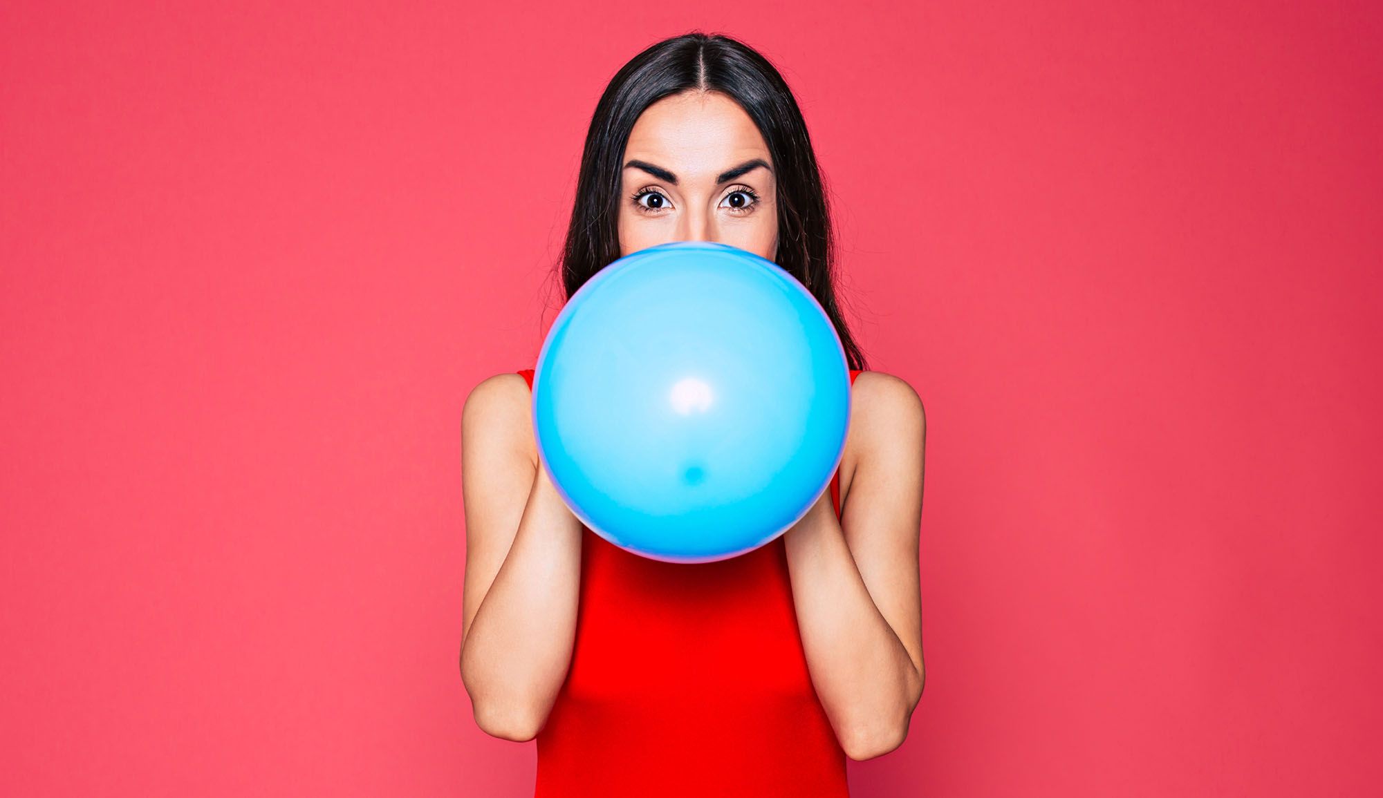 Frau in rotem Kleid, die einen hellblauen Ballon aufbläst vor rotem Hintergrund