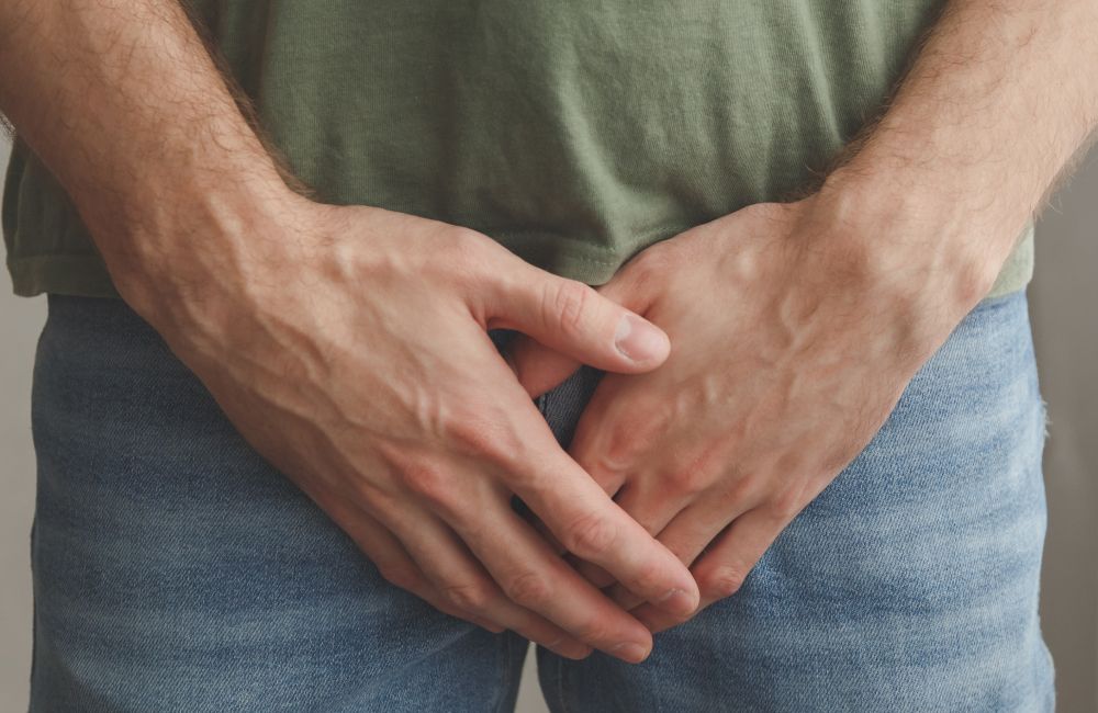 Ein Mann in Jeans und grünem T-Shirt hält seine Hände schützend vor seinen Unterleib