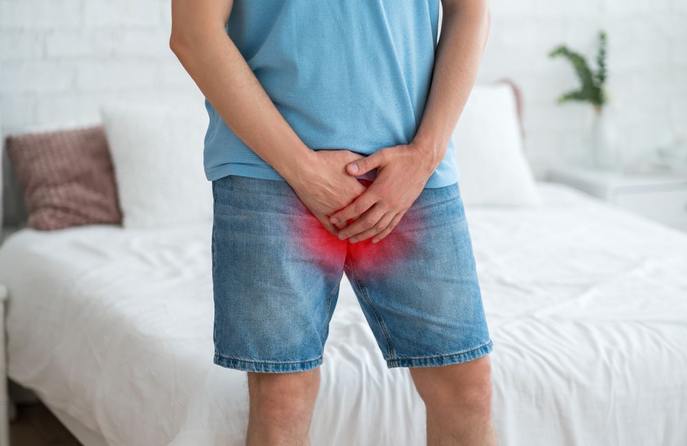 Mann in Jeansshorts und blauem T-Shirt hält sich den schmerzenden Unterleib, der rot markiert ist.