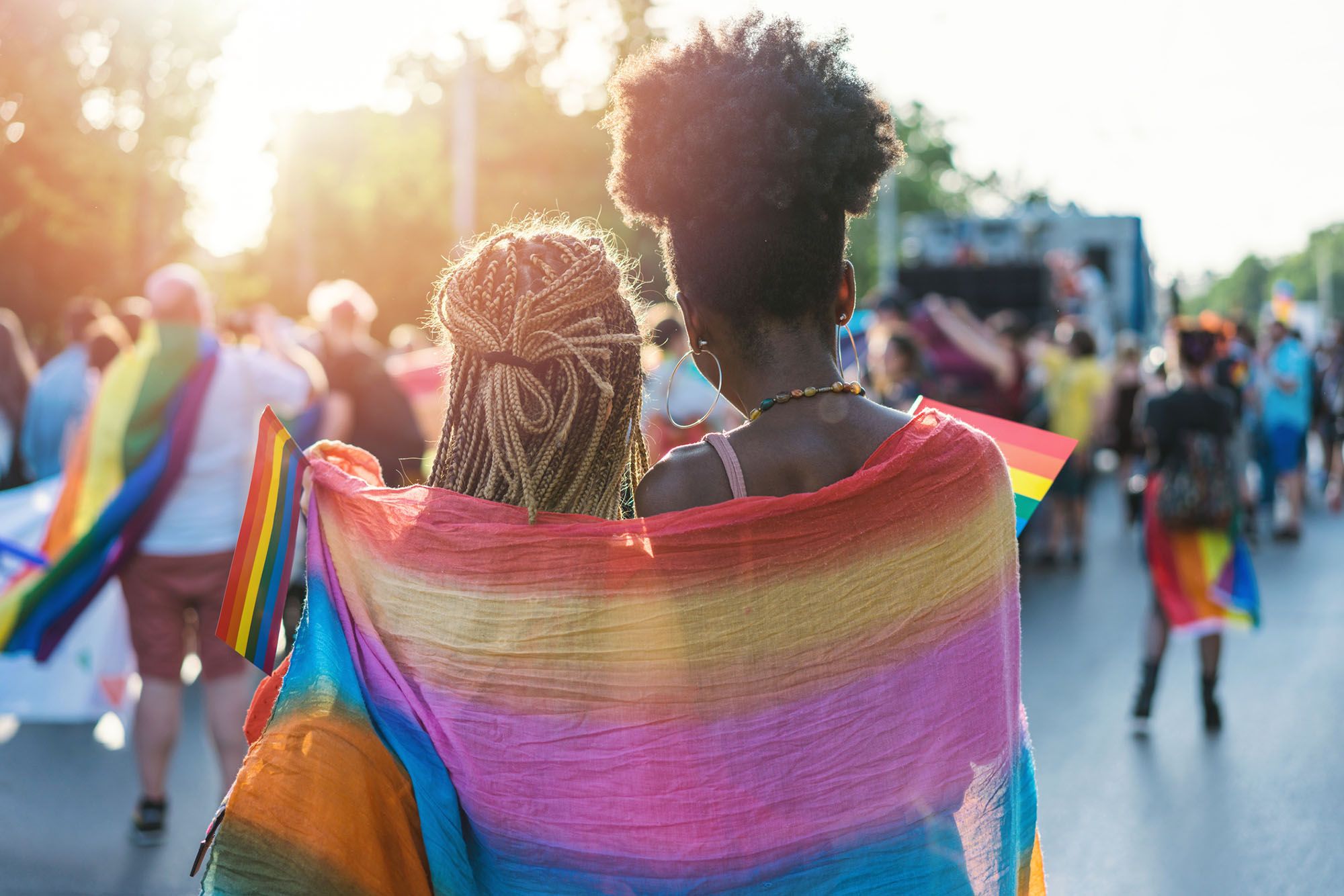 Zwei Frauen stehen während einer Pride-Parade eng beieinander und haben eine Regenbogen-Flagge um sich gelegt.