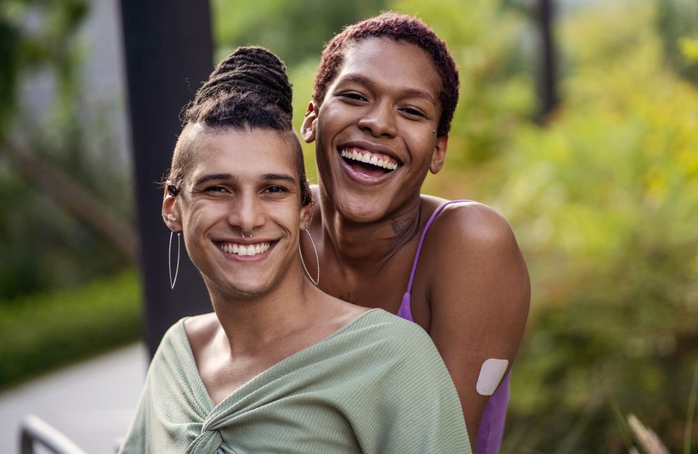 Zwei queere Personen stehen lächelnd nah hintereinander.