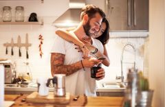 Ein Paar steht in einer Küche und schaut gemeinsam in ein Handy. Die Frau steht hinter dem Mann und legt die Arme um ihn.