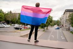 Eine Person spannt eine bi-Flagge hinter sich auf und steht erhöht gut sichtbar über einer Straße