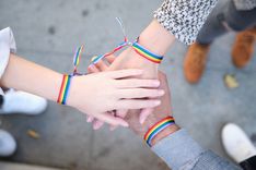 Drei Hände sind in der Mitte des Bildes übereinander gelegt. Jede Hand trägt ein Regenbogenarmband.