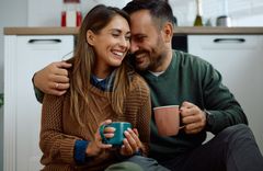 Frau in blauem Hemd und braunem Pullover mit einer Tasse in der Hand, die von einem Mann in grünem Pullover und Tasse in der Hand seitlich umarmt wird während beide auf einem Küchenboden sitzen