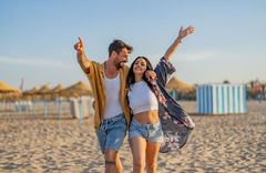 Frau in Hotpants und weißem Shirt und Blumenbluse und ein Mann in weißem Shirt und blauer Shorts und gelbem Hemd an einem Strand